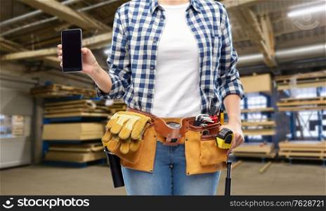 repair, construction and building concept - female worker with smartphone and working tools on belt over workshop at woodworking factory background. female worker with phone and tools at factory