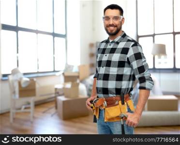 repair, construction and building concept - happy smiling male worker or builder in goggles with tool belt over home room background. happy male builder with tool belt at home