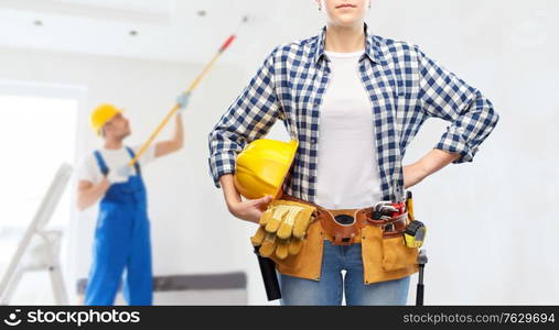 repair, construction and building concept - woman or builder with helmet and working tools on belt over painter painting ceiling background. woman or builder with helmet and working tools