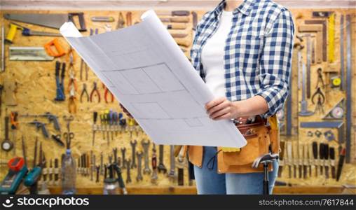 repair, construction and building concept - woman or builder with helmet, blueprint and working tools on belt over workshop on background. female builder with blueprint and working tools