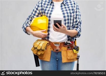 repair, construction and building concept - woman or builder with smartphone, helmet and working tools on belt over grey background. woman or builder with phone and working tools