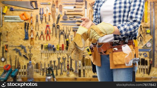 repair, construction and building concept - woman or builder with working tools on belt putting protective gloves on over workshop on background. woman or builder with gloves and working tools