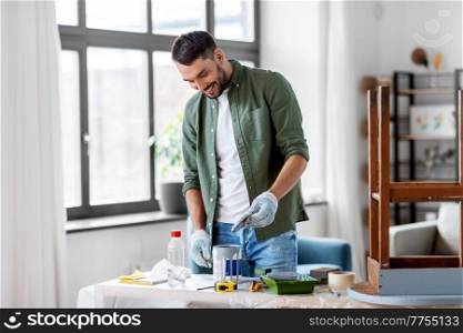 repair, diy and home improvement concept - happy smiling man in protective gloves opening can with grey color paint for painting old wooden table. man opening can with grey color at home