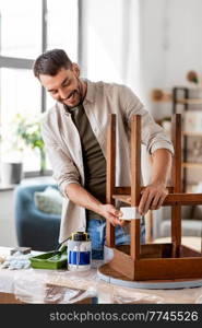 repair, diy and home improvement concept - happy smiling man sticking adhesive tape to old wooden table for repainting it. man sticking adhesive tape to table for repainting