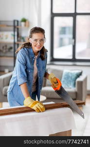 repair, diy and home improvement concept - happy smiling woman in goggles with saw sawing wooden board. woman with saw sawing wooden board at home