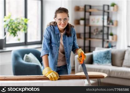 repair, diy and home improvement concept - happy smiling woman in goggles with saw sawing wooden board. woman with saw sawing wooden board at home