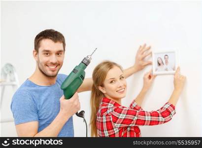 repair, interior design, building, renovation and home concept - smiling couple drilling hole in wall and putting picture up at home
