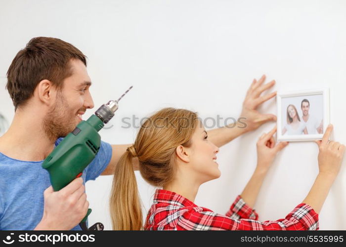 repair, interior design, building, renovation and home concept - smiling couple drilling hole in wall and putting picture up at home