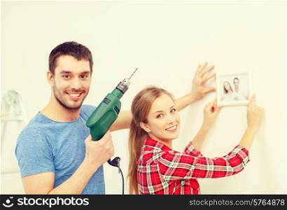 repair, interior design, building, renovation and home concept - smiling couple drilling hole in wall and putting picture up at home