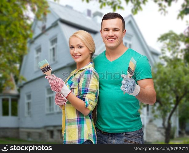 repair, people, real estate, home and family concept - smiling couple with paint brushes over house background