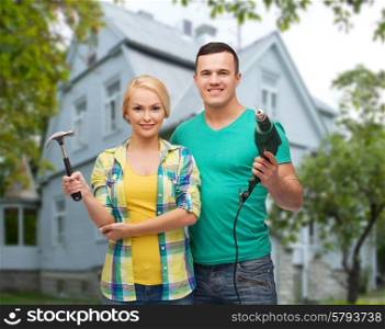 repair, people, real estate, home and family concept - smiling couple with hammer and electric drill over house background