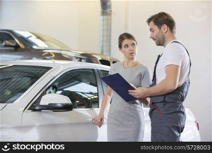 Repairman holding clipboard while conversing with female customer in automobile repair shop
