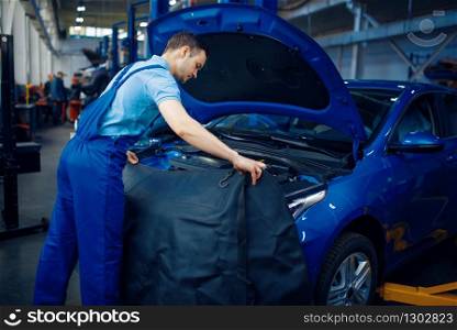 Repairman in uniform checks vehicle engine, car service station. Automobile checking and inspection, professional diagnostics and repair. Repairman checks vehicle engine, car service