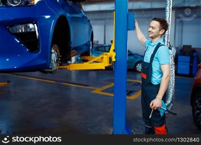Repairman in uniform lifts vehicle on car service station. Automobile checking and inspection, professional diagnostics and repair. Repairman lifts vehicle on car service station