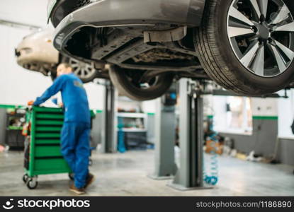 Repairman in uniform near tool box against a car on the lift, suspension diagnostic. Automobile service, vehicle maintenance