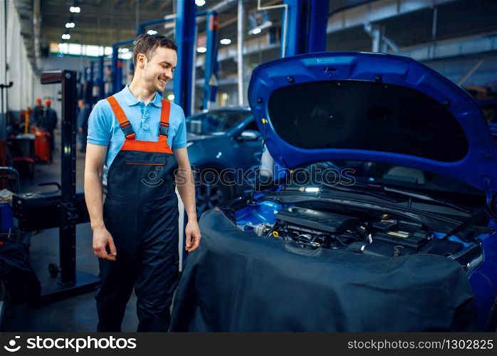 Repairman in uniform stands at vehicle with opened hood, car service station. Automobile checking and inspection, professional diagnostics and repair. Repairman at vehicle with opened hood, car service