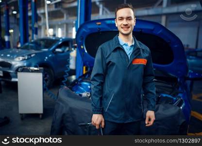 Repairman in uniform stands at vehicle with opened hood, car service station. Automobile checking and inspection, professional diagnostics and repair. Repairman at vehicle with opened hood, car service