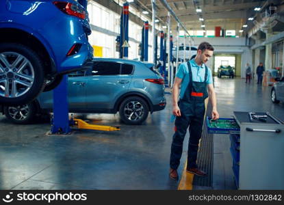 Repairman stands at vehicles on lifts, car service station. Automobile checking and inspection, professional diagnostics and repair. Repairman stands at vehicles on lifts, car service