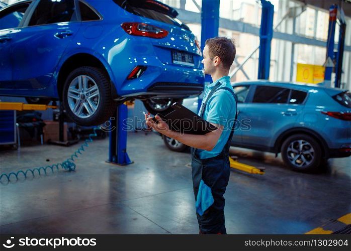 Repairman with a checklist stands at vehicle on lift, car service station. Automobile checking and inspection, professional diagnostics and repair. Repairman with a checklist, car service station