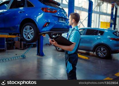 Repairman with a checklist stands at vehicle on lift, car service station. Automobile checking and inspection, professional diagnostics and repair. Repairman with a checklist, car service station