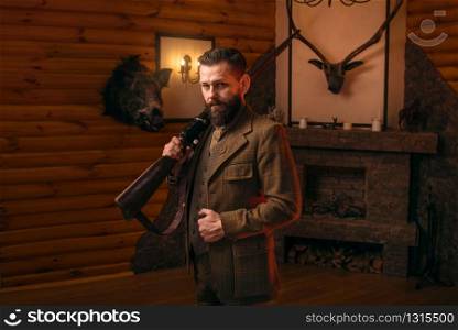 Respectable hunter man in vintage stylish hunting clothing with antique rifle against fireplace. Stuffed wild animals, bear skin and other trophies on background. Hunt lifestyle. Hunter man in vintage clothing with antique rifle