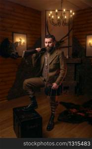 Respectable hunter man with old gun in retrro style traditional hunting clothing standing against antique chest. Retro rifle, vintage clothes. Room with fireplace, stuffed wild animals, bear skin and other trophies on background. Respectable hunter man with old gun