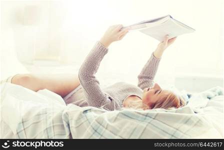 rest, comfort, leisure and people concept - happy young woman reading book in bed at home bedroom. young woman reading book in bed at home