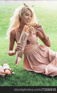 Rest in park, outdoor, people and leisure concept - beauty blonde alone young woman resting in the park. Beauty blonde young woman eating baguette in the park