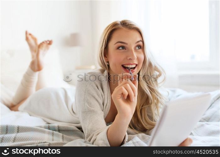rest, inspiration, creativity, comfort and people concept - happy young woman with pen and notebook dreaming in bed at home bedroom