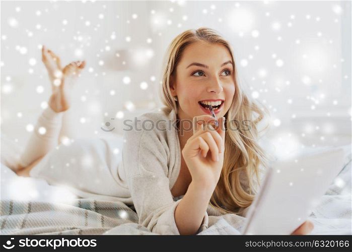 rest, inspiration, creativity, comfort and people concept - happy young woman with pen and notebook dreaming in bed at home bedroom over snow. happy young woman with notebook in bed at home