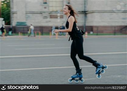 Rest leisure and lifestyle concept. Cheerful dark haired young European woman breathes fresh air while rollerskating has hobby leads active lifestyle poses outdoor engaged in fitness activity
