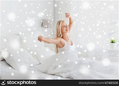 rest, sleeping, comfort and people concept - happy young woman stretching in bed after waking up at home bedroom over snow