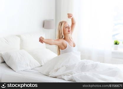 rest, sleeping, comfort and people concept - happy young woman stretching in bed after waking up at home bedroom