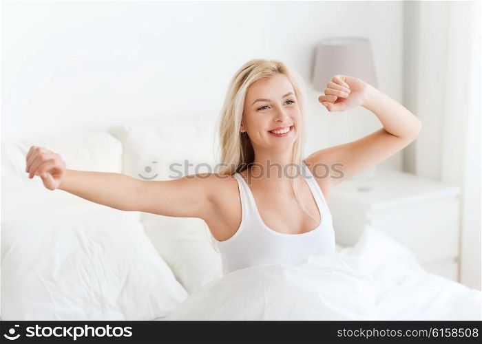 rest, sleeping, comfort and people concept - happy young woman stretching in bed after waking up at home bedroom