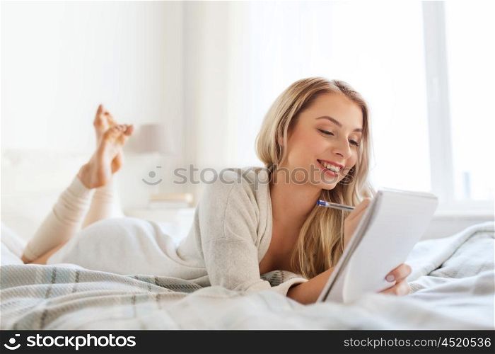 rest, sleeping, comfort and people concept - happy young woman with pen and notebook writing in bed at home bedroom