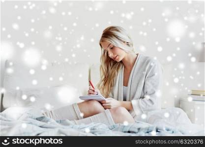 rest, sleeping, comfort and people concept - happy young woman with pen and notebook writing in bed at home bedroom over snow. happy young woman with notebook in bed at home