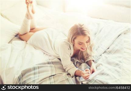 rest, sleeping, comfort and people concept - happy young woman with pen and notebook writing in bed at home bedroom. happy young woman with notebook in bed at home