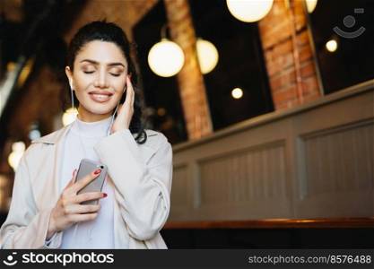 Restful brunette woman with lovely appearance closing her eyes with joy while enjoying loud music in her earphones. Stylish woman listening to her favourite music while having free time and relaxation