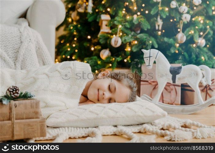 Restful small child lies on soft white pillow, sees pleasant dreams, fall asleep near decorated New Year tree, waits for presents before Christmas Eve, has tender smile on face. Children, coziness