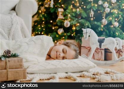 Restful small child lies on soft white pillow, sees pleasant dreams, fall asleep near decorated New Year tree, waits for presents before Christmas Eve, has tender smile on face. Children, coziness
