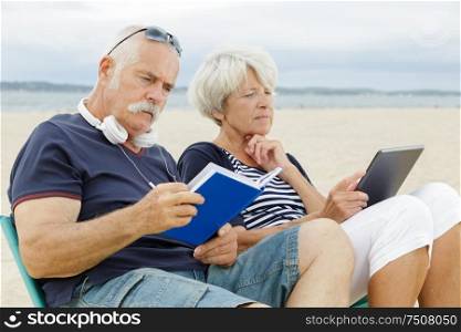 retired couple reading book and tablet on the beach