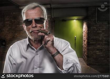 Retired man with strong personality. Retired man with strong personality standing in the darkness in sunglasses puffing on a big cigar, quintessential stereotype of a retired successful executive