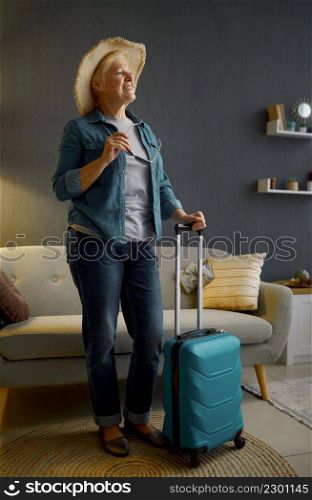 Retired woman in sunhat standing with baggage in home living-room. Preparation for summer vacation. Senior woman in sunhat standing with baggage