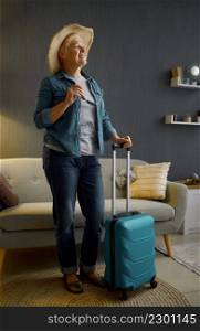 Retired woman in sunhat standing with baggage in home living-room. Preparation for summer vacation. Senior woman in sunhat standing with baggage
