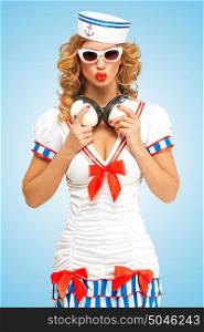 Retro photo of a fashionable pin-up sailor girl in sunglasses with duck lips, holding big vintage music headphones around her neck on blue background.