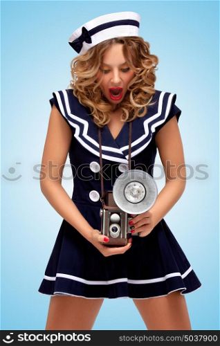 Retro photo of a fashionable pin-up sailor girl looking surprisingly at an old vintage photo camera with bulb flash on blue background.