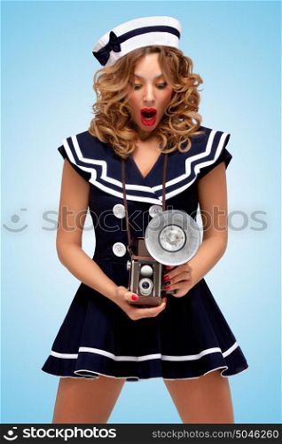 Retro photo of a fashionable pin-up sailor girl looking surprisingly at an old vintage photo camera with bulb flash on blue background.