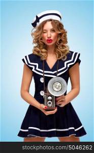 Retro photo of a fashionable pin-up sailor girl with an old vintage photo camera with bulb flash winking to the camera on blue background.