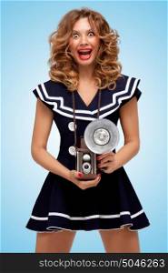 Retro photo of a fashionable pin-up sailor girl with an old vintage photo camera with bulb flash winking to the camera on blue background.