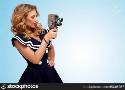 Retro photo of a glamorous pin-up sailor girl with an old vintage cinema 8 mm camera, looking like a sexy producer, shooting a movie on blue background.